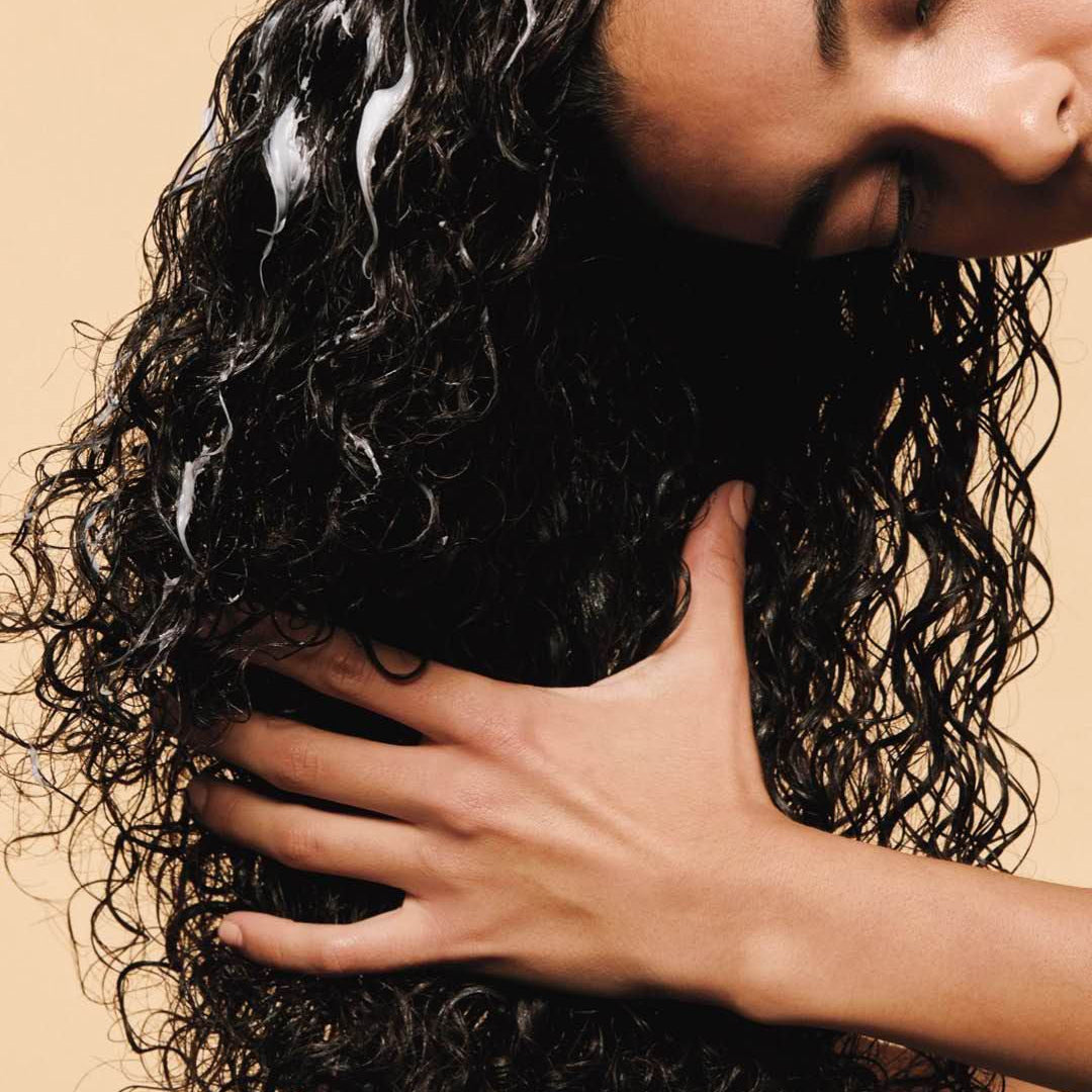 Close-up of a woman with wet, dark curly hair and white cream distributed through the curls, gently holding her hair.