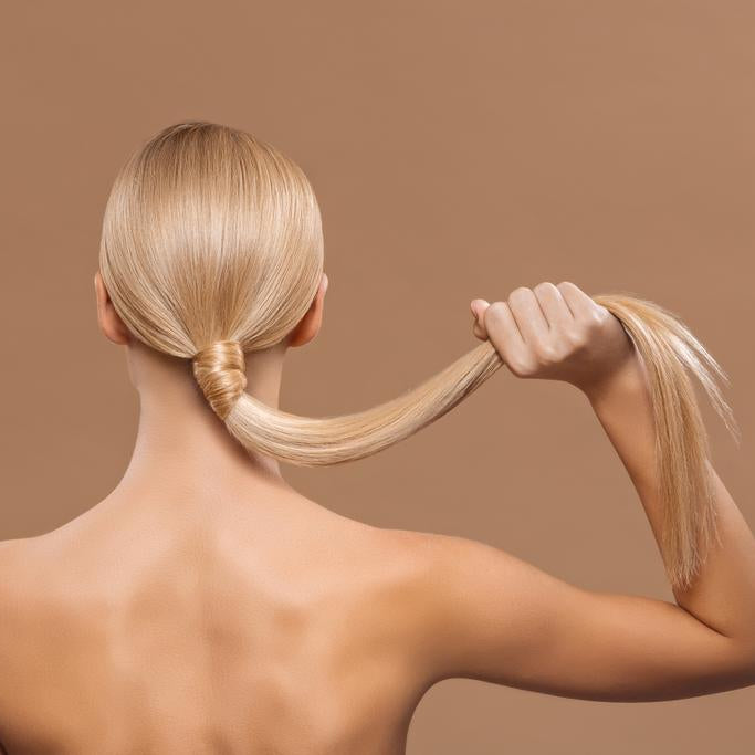 Back view of a woman holding her sleek blonde ponytail against a warm neutral background, showcasing smooth, glossy hair.