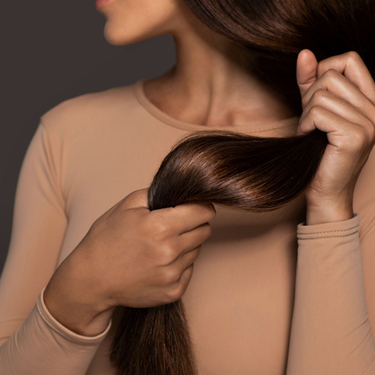 Woman in a beige top holding a section of her long, glossy brown hair against a dark background.